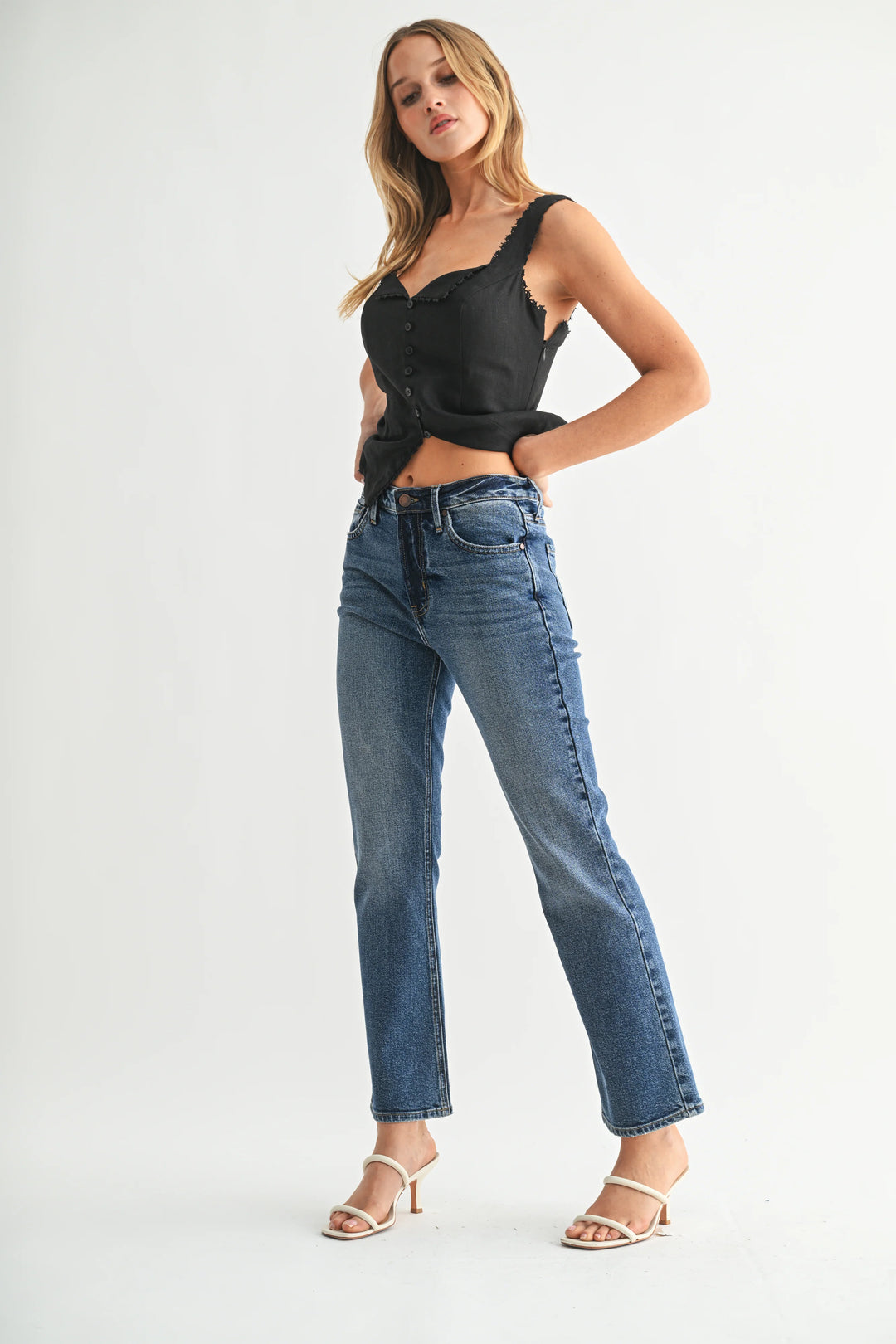 A woman modeling a black lace-trimmed sleeveless top and high-waisted blue jeans with white heeled sandals, standing against a light gray studio background.
