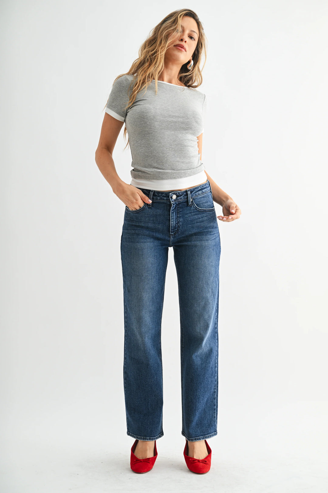 A person wearing medium-wash blue jeans with a straight-leg fit, paired with a gray top and red flats, standing against a light gray studio background.