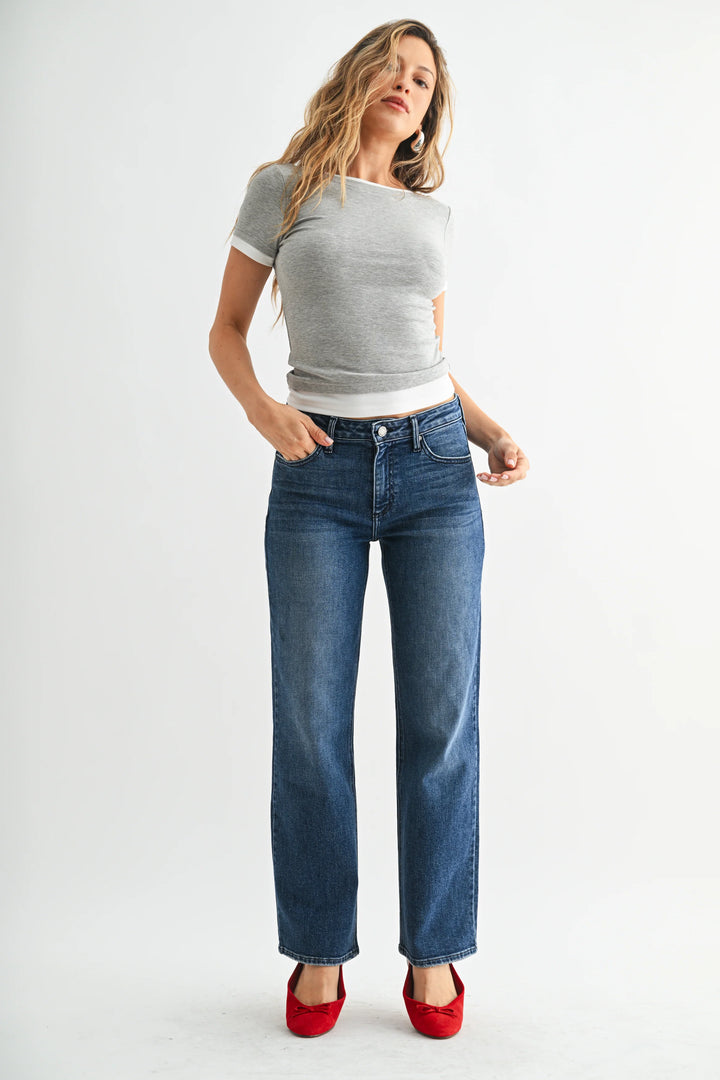 A person wearing medium-wash blue jeans with a straight-leg fit, paired with a gray top and red flats, standing against a light gray studio background.