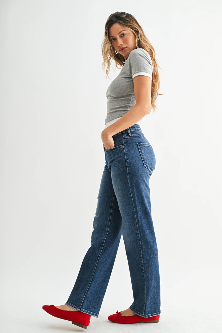 A person wearing medium-wash blue jeans with a straight-leg fit, paired with a gray top and red flats, standing against a light gray studio background.