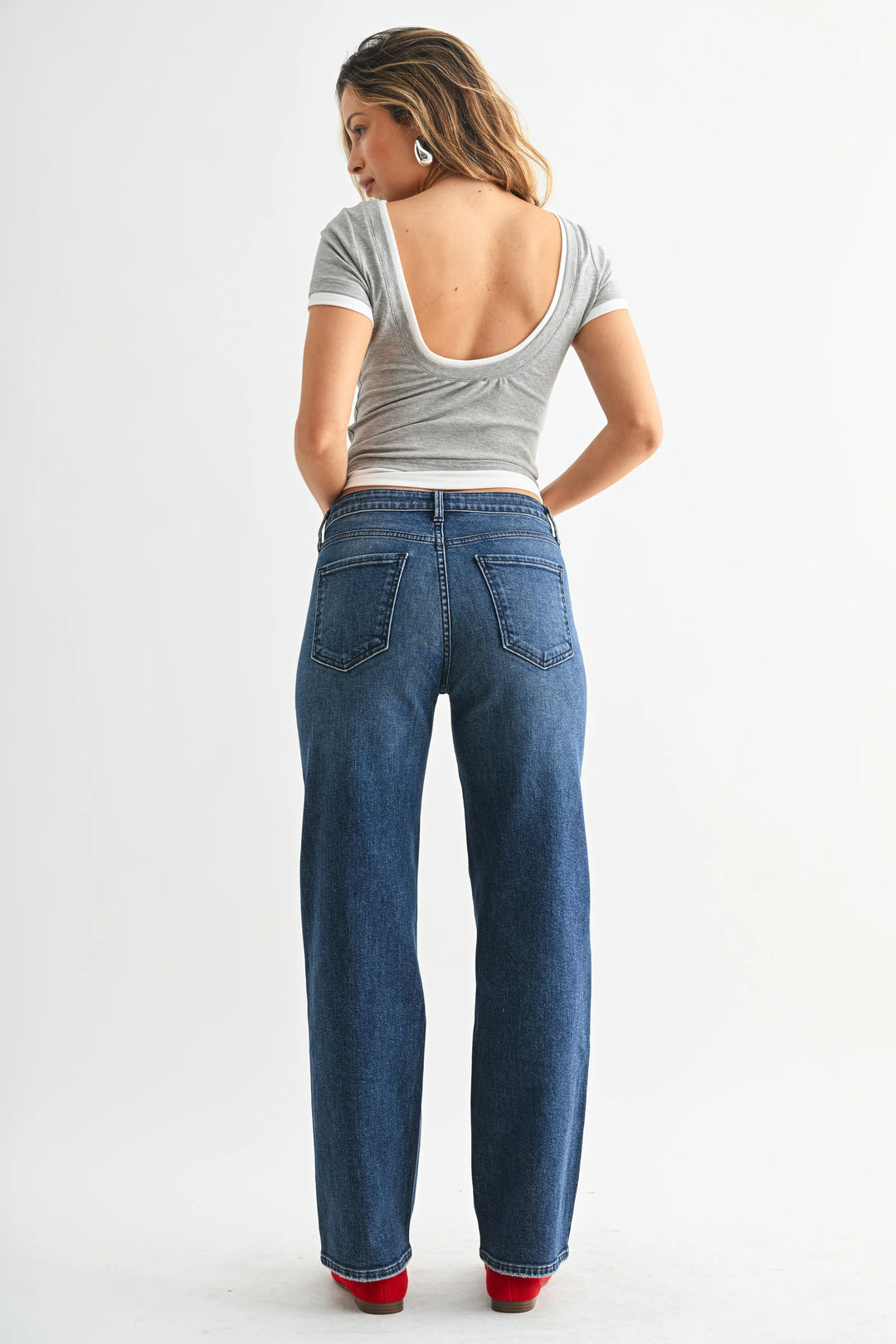 A person wearing medium-wash blue jeans with a straight-leg fit, paired with a gray top and red flats, standing against a light gray studio background.
