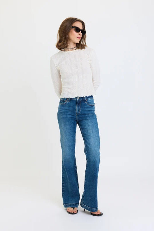 A person wearing medium-wash blue flare jeans with a white ribbed long-sleeve top and black flip-flop sandals, standing against a plain white studio background.