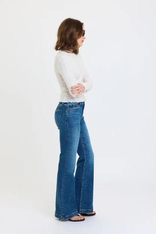 A person wearing medium-wash blue flare jeans with a white ribbed long-sleeve top and black flip-flop sandals, standing against a plain white studio background.