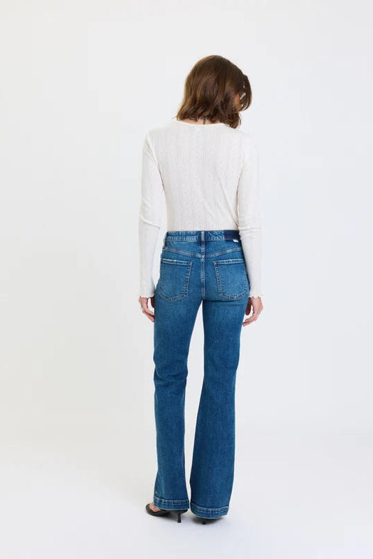 A person wearing medium-wash blue flare jeans with a white ribbed long-sleeve top and black flip-flop sandals, standing against a plain white studio background.
