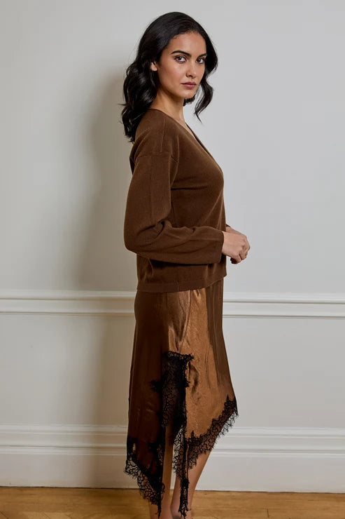 A woman stands in profile wearing a brown long-sleeve knit top and a matching brown satin slip dress with black lace trim, against a plain off-white wall.
