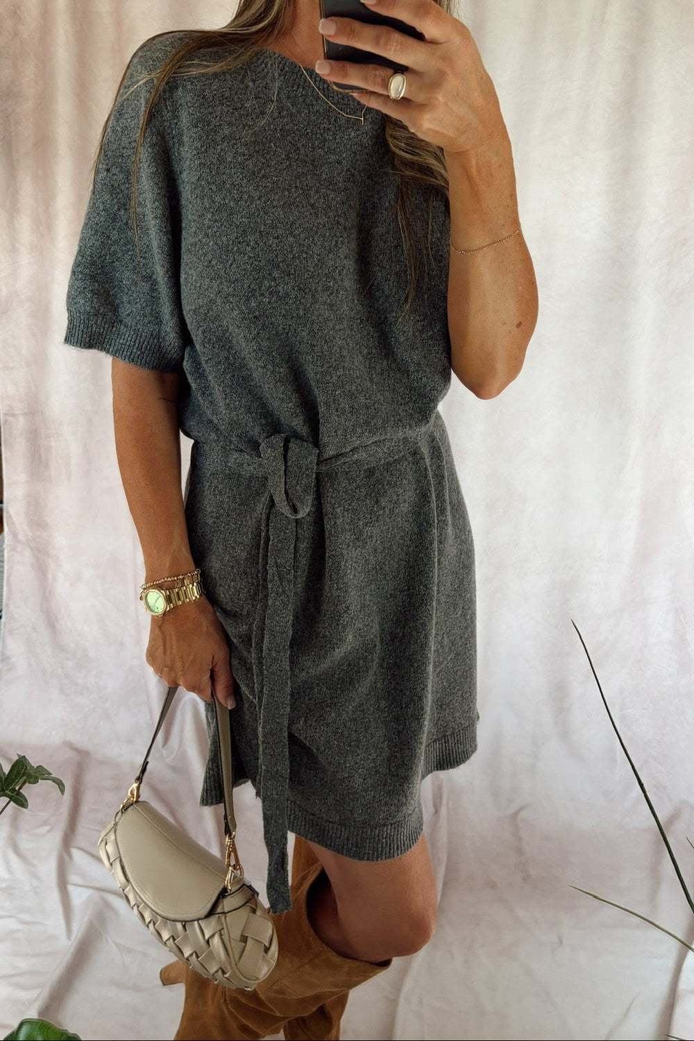 A woman takes a mirror selfie wearing a dark gray short-sleeve knit romper with a tie waist, paired with brown boots and a cream-colored handbag, set against a draped white fabric backdrop.