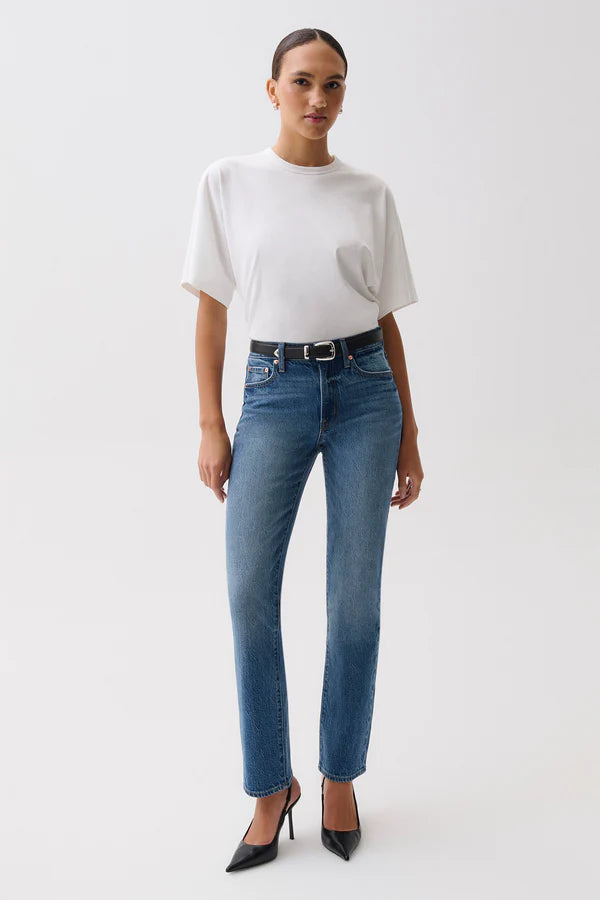 A woman modeling a pair of blue straight-leg jeans with a black leather belt, paired with a white t-shirt, against a plain light gray studio background. pistola fall 2026