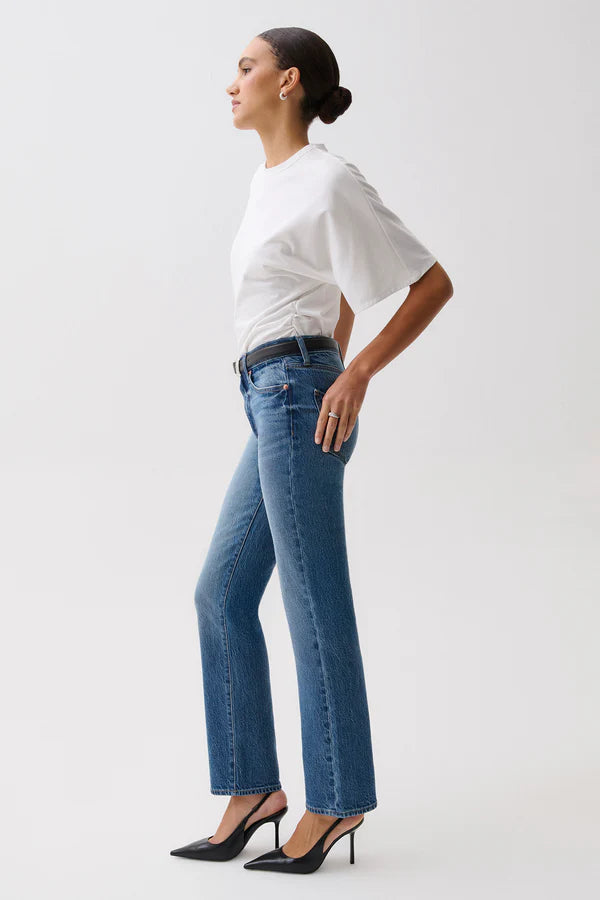 A woman modeling a pair of blue straight-leg jeans with a black leather belt, paired with a white t-shirt, against a plain light gray studio background. pistola fall 2026