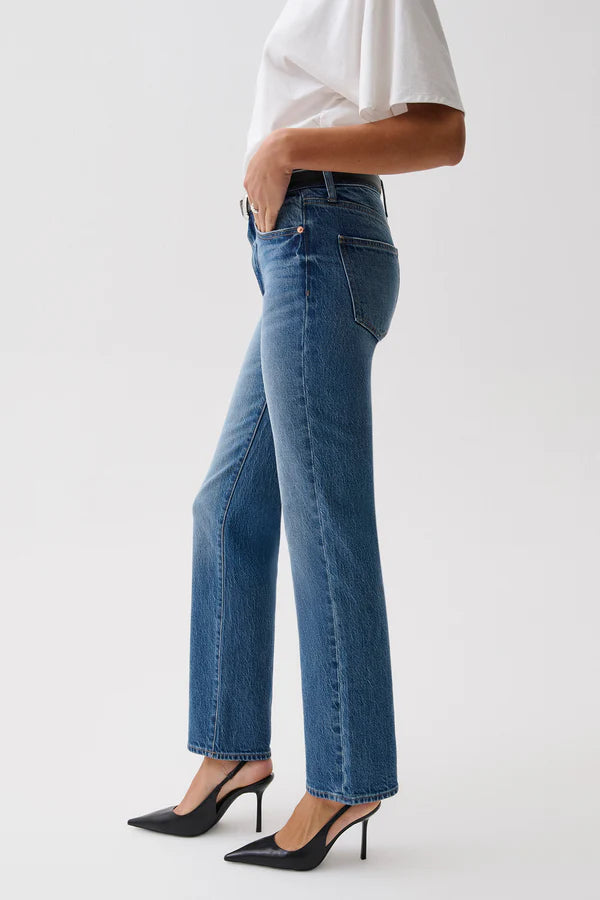 A woman modeling a pair of blue straight-leg jeans with a black leather belt, paired with a white t-shirt, against a plain light gray studio background. pistola fall 2026
