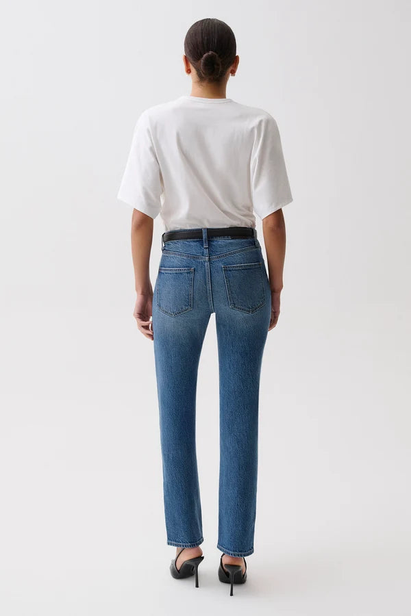A woman modeling a pair of blue straight-leg jeans with a black leather belt, paired with a white t-shirt, against a plain light gray studio background. pistola fall 2026