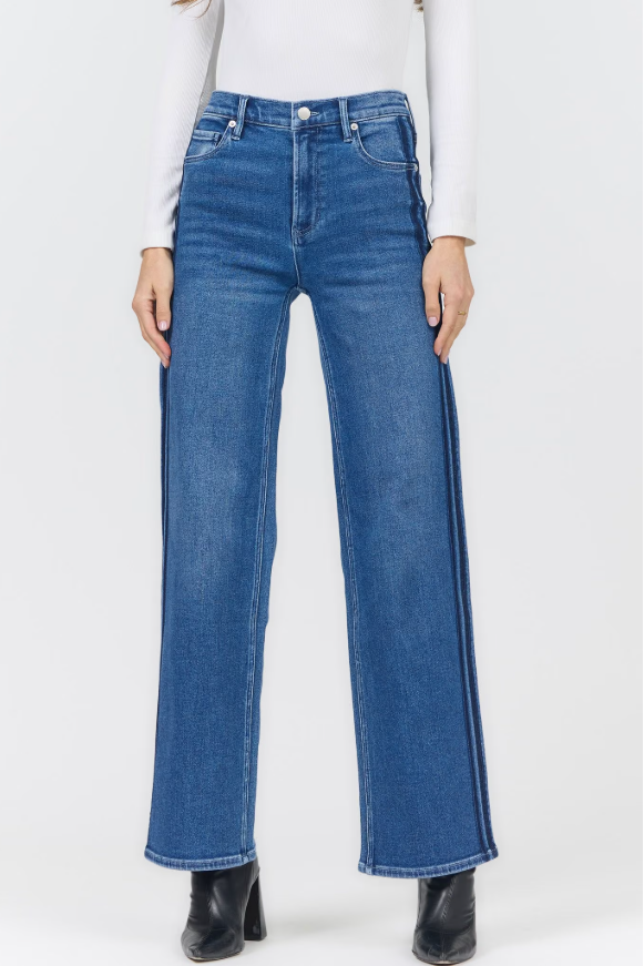 A model wearing blue wide-leg jeans with distinctive vertical white stripes down the outer sides, styled with a white long-sleeve top and black ankle boots against a light gray studio background.