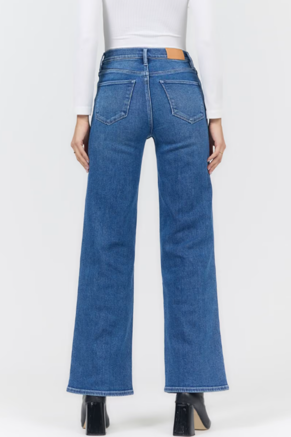 Rear view of a model wearing blue wide-leg jeans with a white long-sleeve top and black heels, shown against a plain light gray studio background.