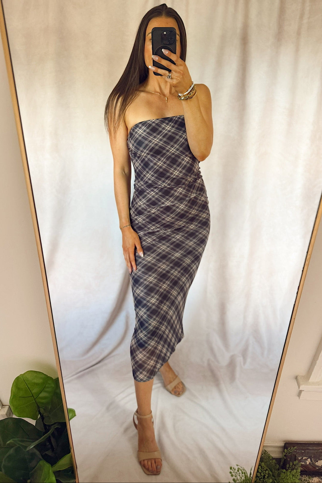A woman takes a mirror selfie wearing a strapless plaid midi dress in gray, white, and navy tones, paired with beige high-heeled sandals.