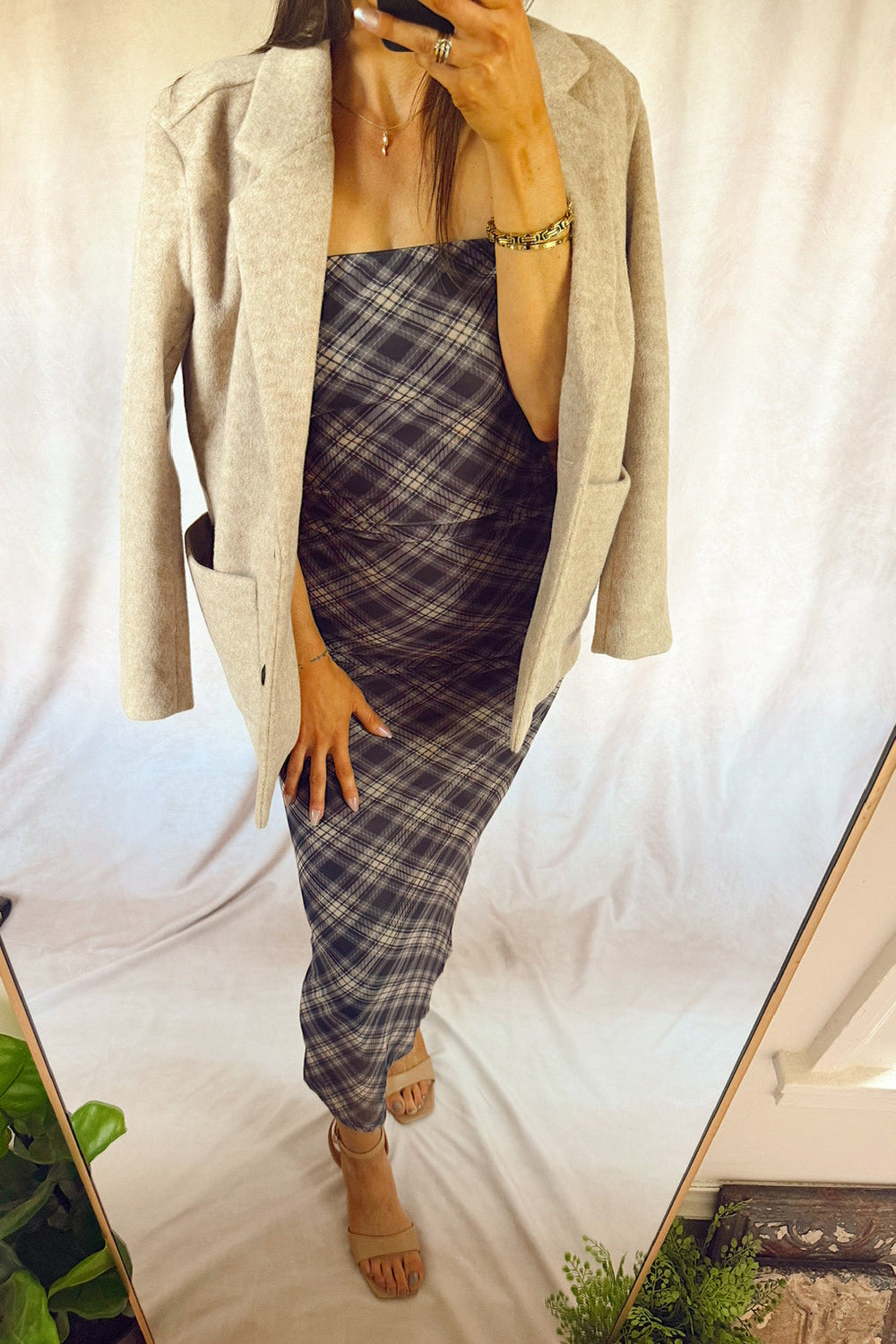 A woman takes a mirror selfie wearing a light beige open-front blazer layered over a dark gray and white plaid strapless jumpsuit, paired with beige heeled sandals.