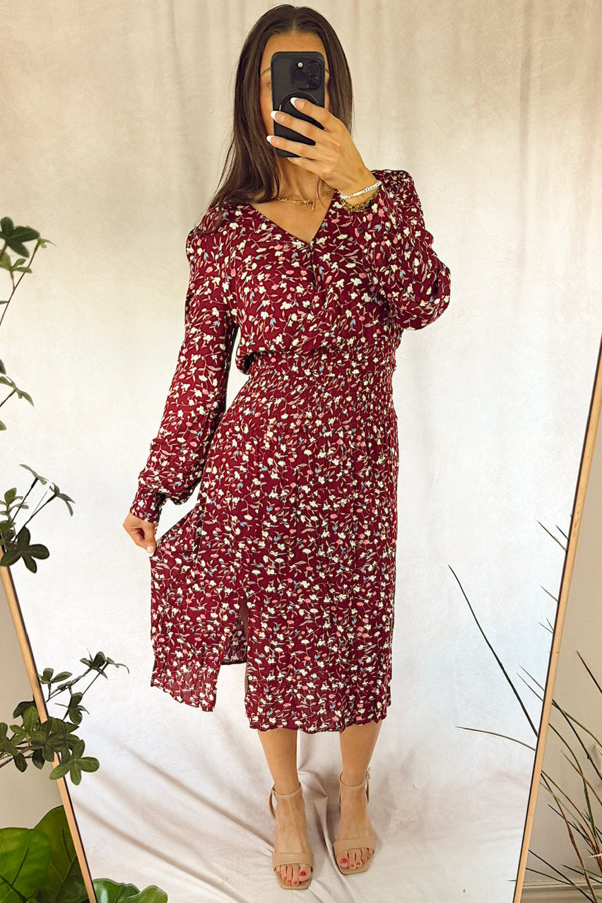 A woman takes a mirror selfie wearing a long-sleeve red midi dress with a white floral pattern, paired with beige high-heeled sandals. The background is a white draped fabric backdrop with a wooden mirror frame and green plants in the foreground.