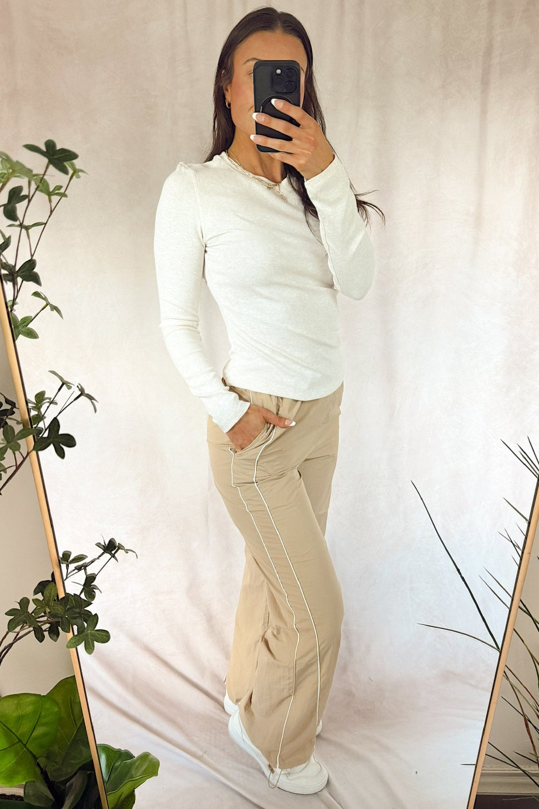 A woman takes a mirror selfie wearing a white long-sleeve Henley shirt and beige wide-leg cargo pants with white side stripes, set against a draped beige fabric backdrop with indoor plants.