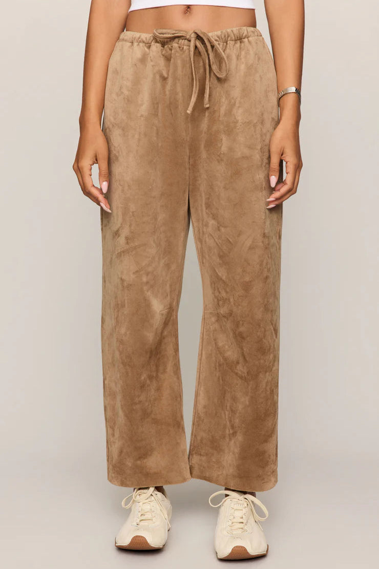 Person wearing tan faux suede pants with offwhite sneakers and a white tank on a plain background