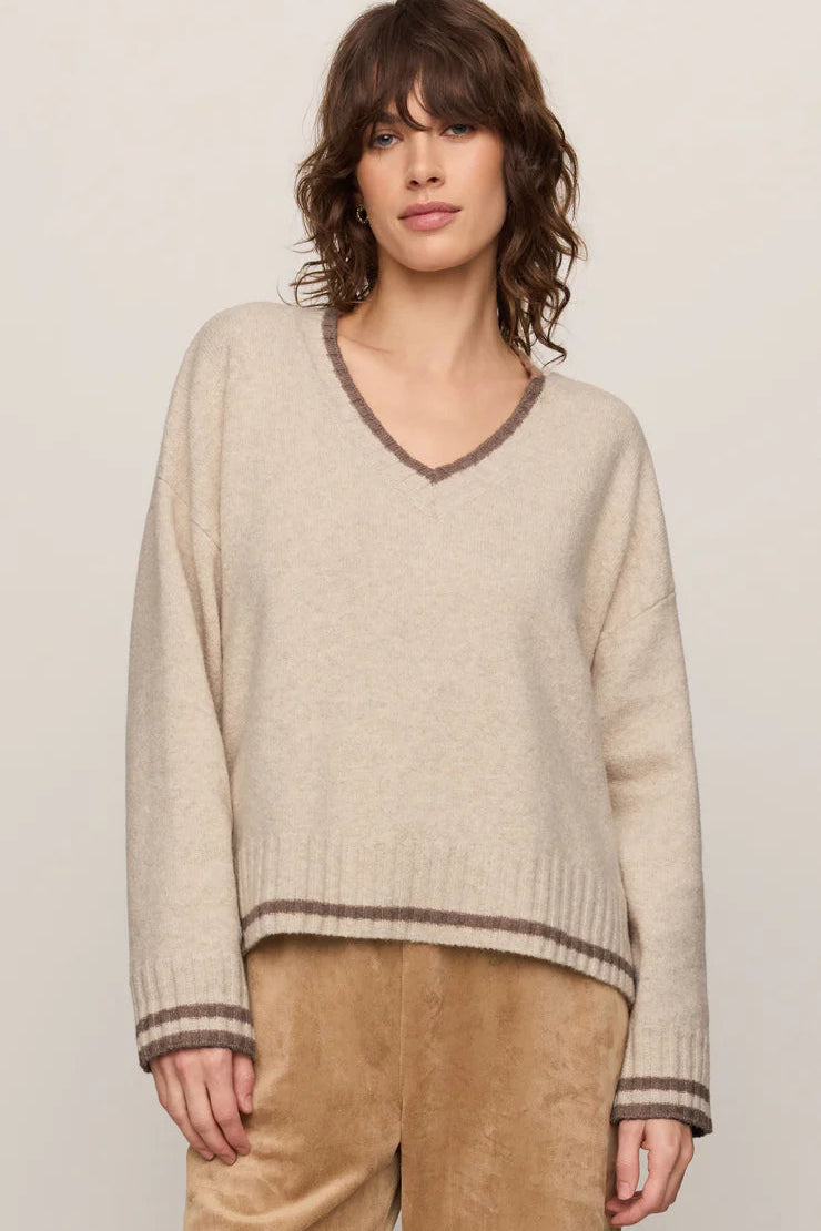A woman wearing a light beige knit sweater with brown trim on the V-neck, cuffs, and hem, paired with tan pants, against a plain light beige background.