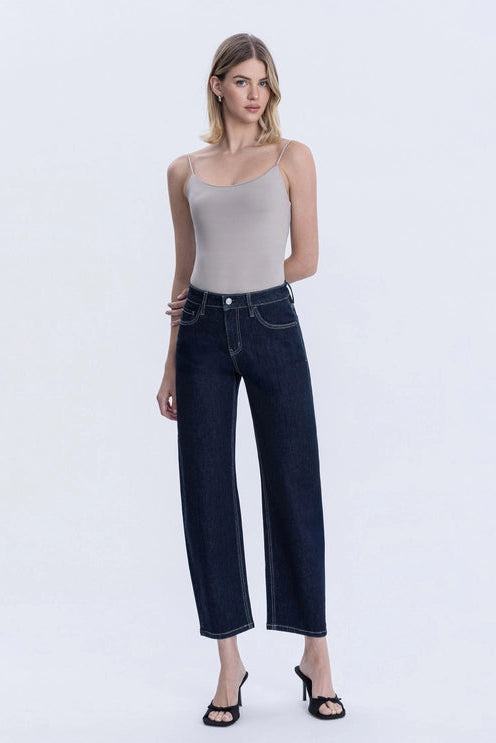 A model wearing dark indigo barrel leg cropped jeans, paired with a light beige top and black strappy sandals, shown against a plain white studio background.