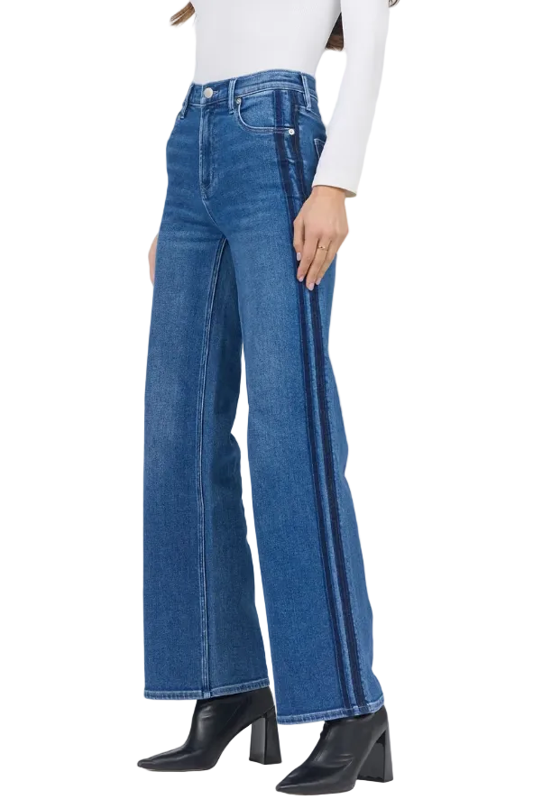 A model wearing blue wide-leg jeans with dark vertical stripe detailing down the outer sides, styled with a white long-sleeve top and black pointed-toe heeled boots against a light gray studio background.