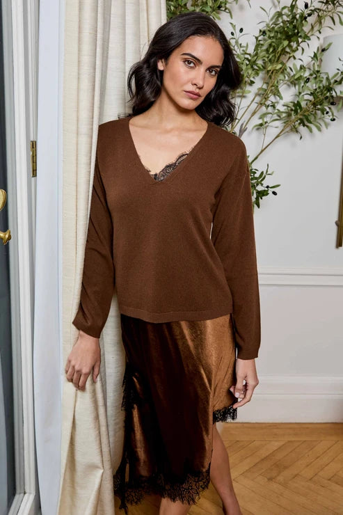 A woman models a brown knit long-sleeve top with a lace-trimmed V-neck, paired with a matching brown satin slip skirt with lace trim, in an indoor setting with a cream curtain and wooden floor.