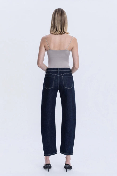 A model wearing dark indigo barrel leg cropped jeans, paired with a light beige top and black strappy sandals, shown against a plain white studio background.