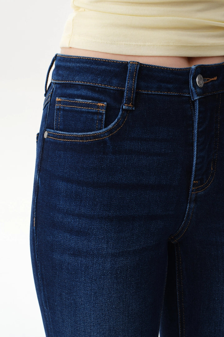 A person wearing dark blue straight-leg  bootcut jeans with a light yellow top and brown sandals, shown from the waist down against a plain white studio background.