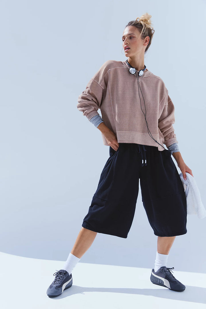 A woman modeling a light brown cropped sweatshirt, black wide-leg shorts, white socks, and dark blue sneakers against a light blue studio background.