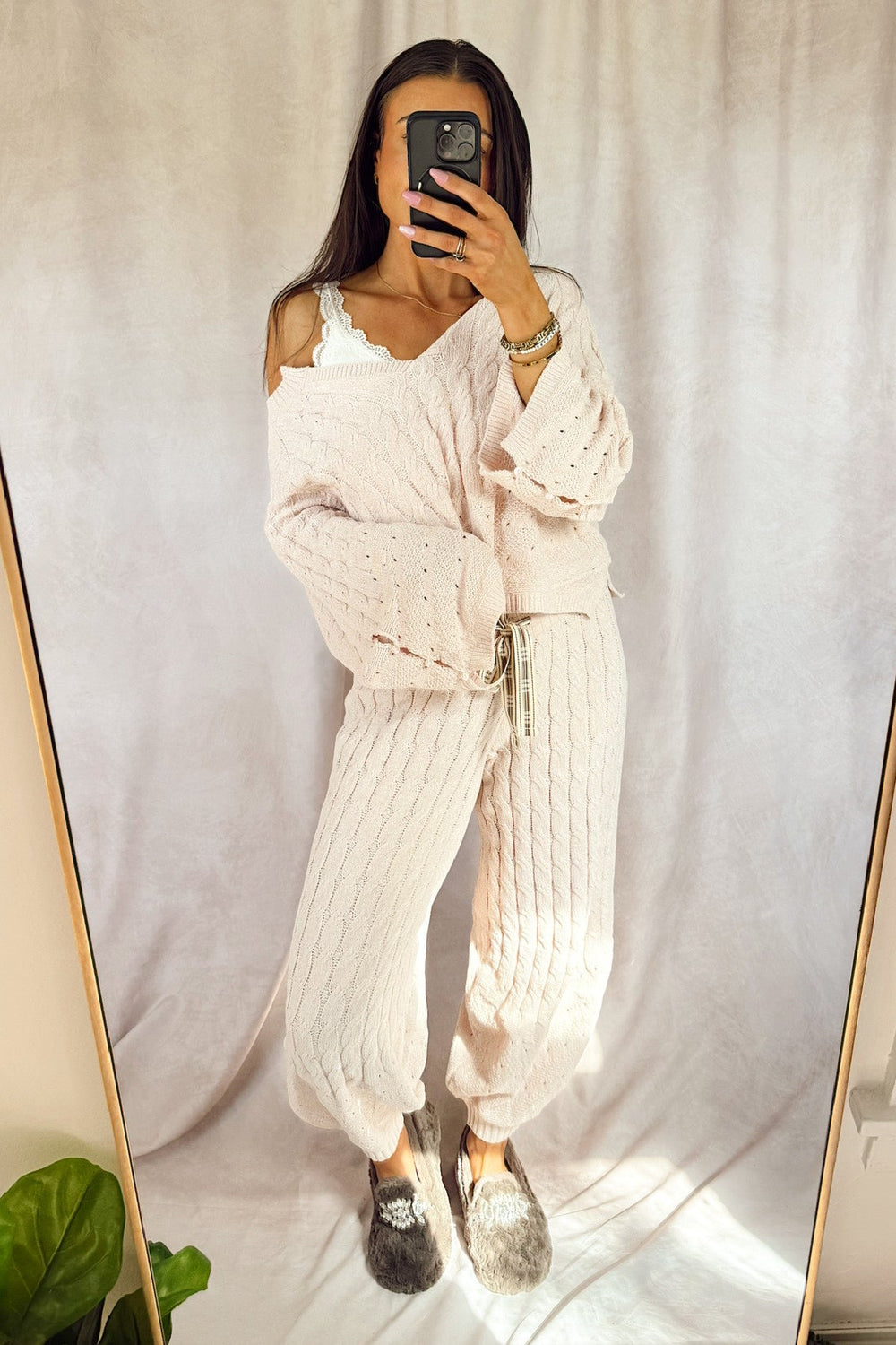 A woman takes a mirror selfie wearing a light pink knit lounge set consisting of a long-sleeve top and matching wide-leg pants, paired with gray slip-on shoes, set against a neutral draped fabric backdrop.