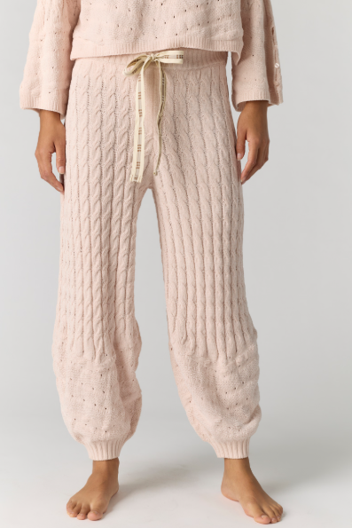 A person wearing light blush pink cable-knit lounge pants with a cream drawstring and a matching textured knit top, shown from the chest down against a light gray studio background.