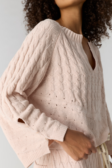 A close-up of a person wearing a light blush pink cable-knit sweater with a V-neckline and long sleeves, shown against a plain light gray background.