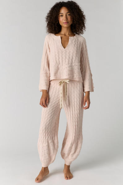 A woman modeling a light pink cable-knit two-piece loungewear set, consisting of a cropped long-sleeve top with a split neckline and matching drawstring harem pants, against a plain light gray studio background.