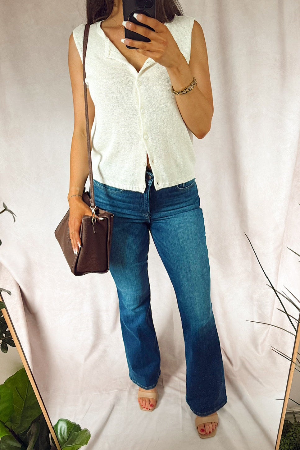 A woman takes a mirror selfie showing an outfit consisting of a sleeveless white knit button-up shirt and blue flare jeans, holding a brown handbag. The background is a draped white fabric with indoor plants visible around the edges. good american. z supply