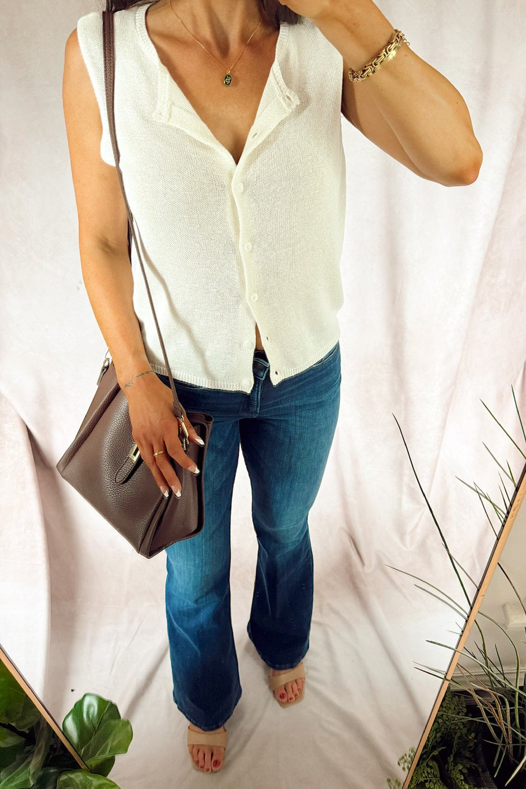 A woman wearing a sleeveless white button-front top and blue jeans, holding a brown shoulder bag, photographed in a mirror selfie against a light draped backdrop with plants visible. good american. z supply.