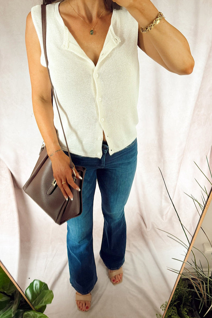 A woman wearing a sleeveless white button-front top and blue jeans, holding a brown shoulder bag, photographed in a mirror selfie against a light draped backdrop with plants visible. good american. z supply.