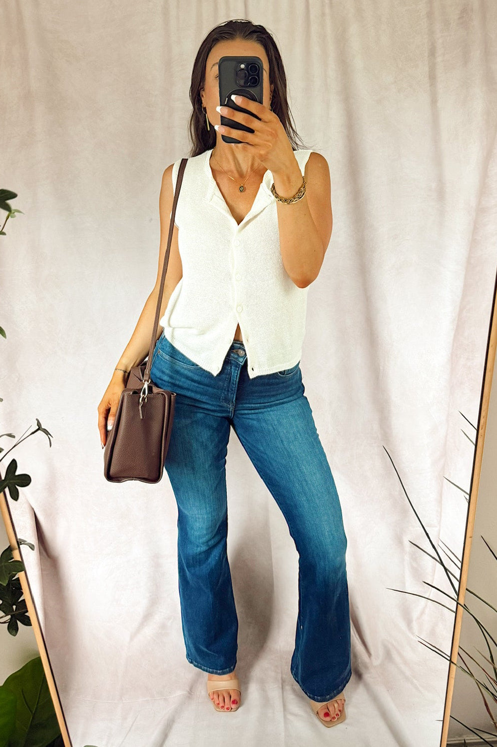 A woman takes a mirror selfie wearing a white sleeveless button-front top and blue flare jeans, holding a brown handbag. The photo is taken indoors in front of a light fabric backdrop with plants visible at the edges. good american. z supply