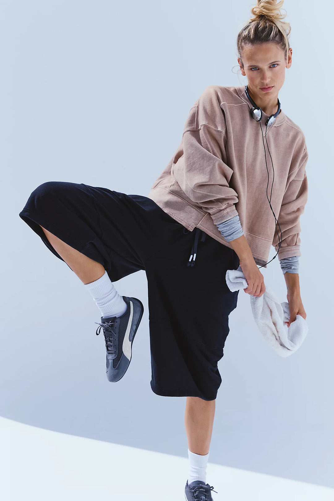 A woman modeling a light brown cropped sweatshirt, black wide-leg shorts, white socks, and dark blue sneakers against a light blue studio background.