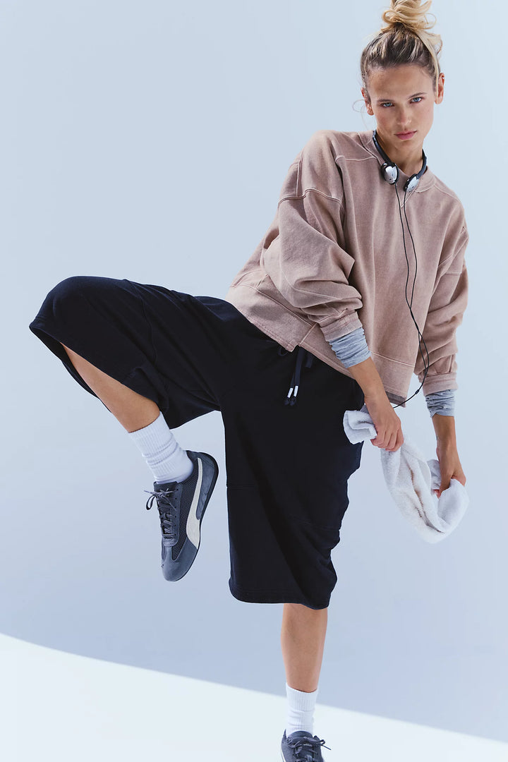 A woman modeling a light brown cropped sweatshirt, black wide-leg shorts, white socks, and dark blue sneakers against a light blue studio background.