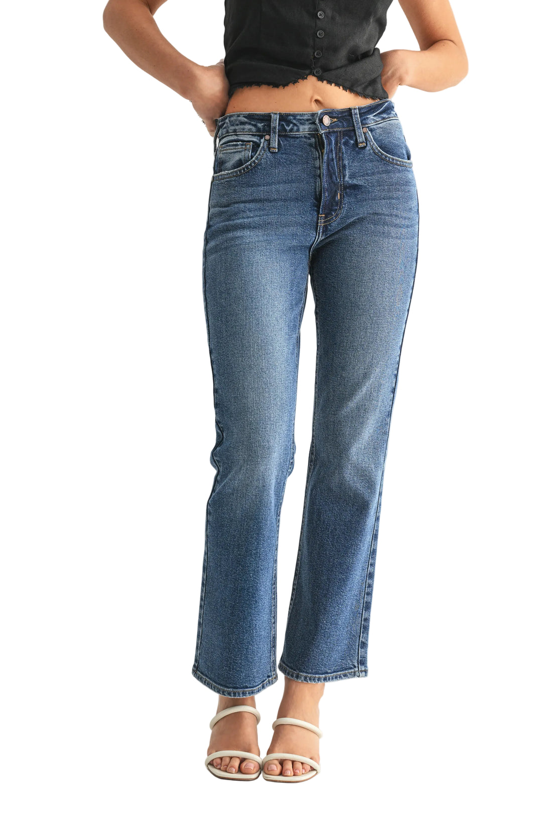 A person wearing medium-wash blue straight-leg jeans with a cropped ankle length, paired with a black top and white heeled sandals, shown from the waist down against a light gray studio background.