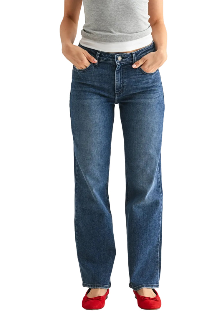 A person wearing medium-wash blue jeans with a straight-leg fit, paired with a gray top and red flats, standing against a light gray studio background.