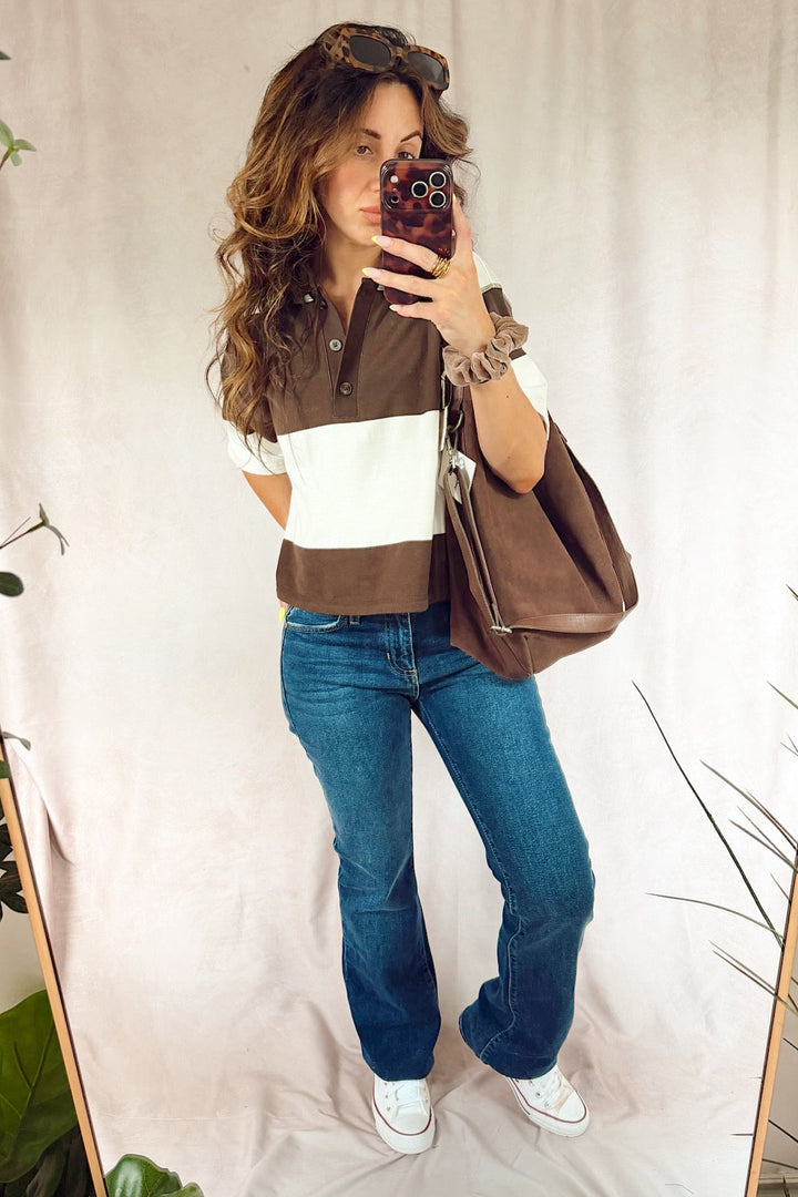 A woman takes a mirror selfie wearing a brown and white color-block short-sleeve polo shirt, blue flared jeans, white sneakers, and carrying a brown leather tote bag. The background is a draped beige fabric backdrop with decorative plants on the sides.