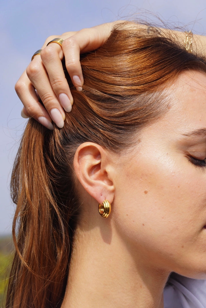 A close-up lifestyle shot of a woman with reddish-brown hair wearing a small gold hoop earring, with her hand resting on her hair. The background shows a soft-focus outdoor sky. may martin cassidy 14k gold dipped hoop earrings