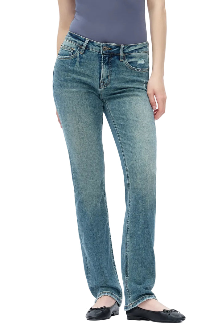 A model wearing a pair of medium-wash blue straight-leg jeans, paired with a slate purple top and black flat shoes, photographed against a seamless white studio background.
