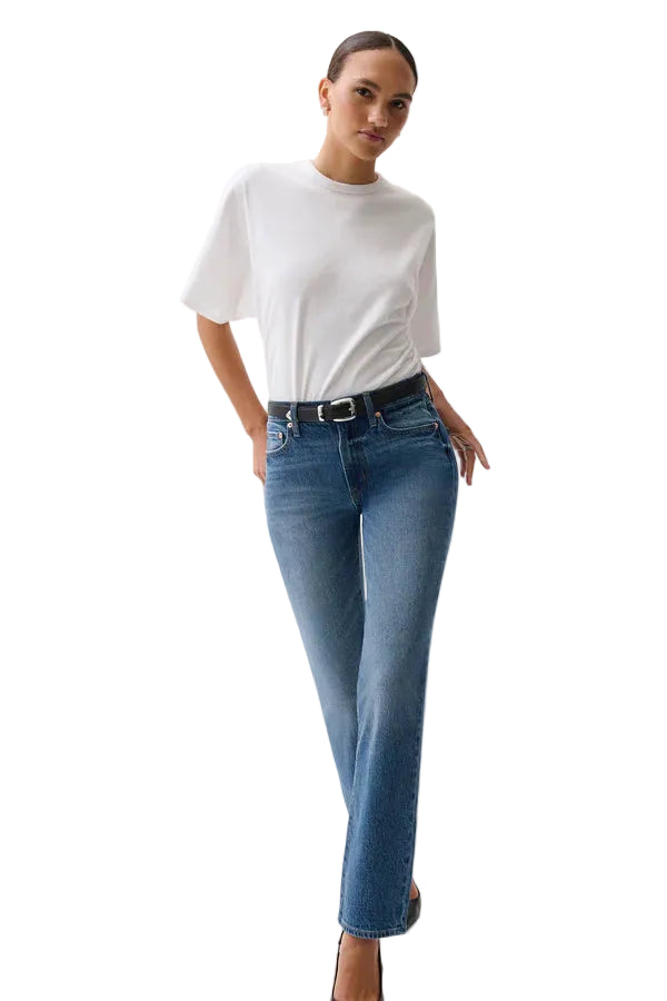 A woman modeling a pair of blue straight-leg jeans with a black leather belt, paired with a white t-shirt, against a plain light gray studio background. pistola fall 2026