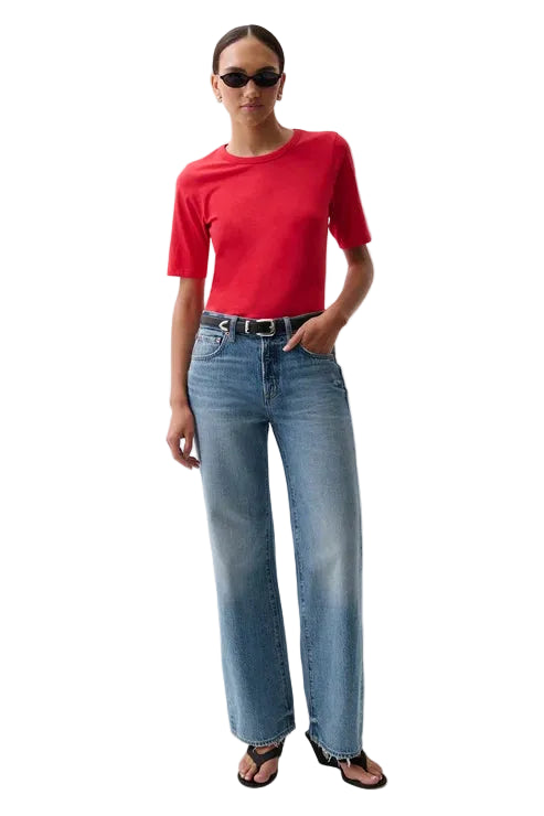 A model wearing a bright red short-sleeve crewneck t-shirt, light-wash blue pistola baggy wide-leg jeans, a black belt, and black sandals, standing against a plain white studio background