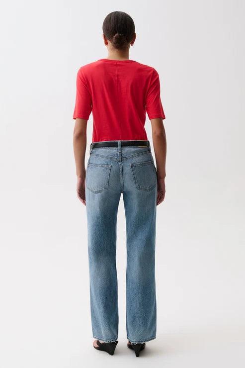 A model wearing a bright red short-sleeve crewneck t-shirt, light-wash blue pistola baggy wide-leg jeans, a black belt, and black sandals, standing against a plain white studio background