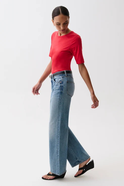 A model wearing a bright red short-sleeve crewneck t-shirt, light-wash blue pistola baggy wide-leg jeans, a black belt, and black sandals, standing against a plain white studio background