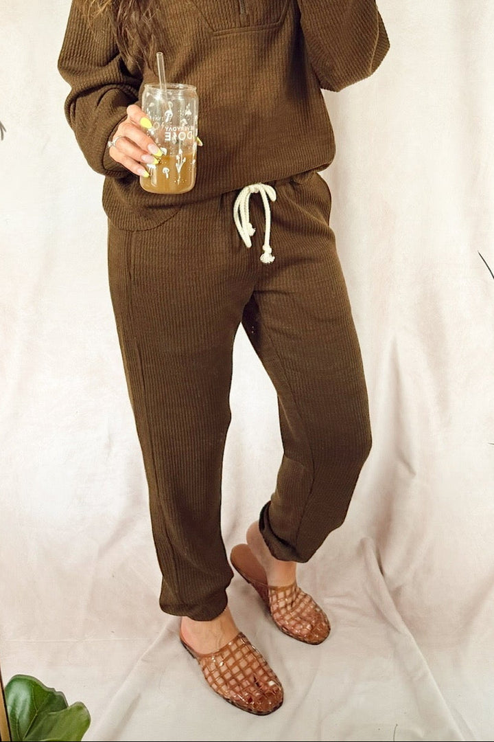 A woman takes a mirror selfie wearing a matching brown two-piece lounge set consisting of a half-zip pullover and jogger pants with a white drawstring. She is holding a glass of iced coffee in front of a white fabric backdrop with plants on either side.