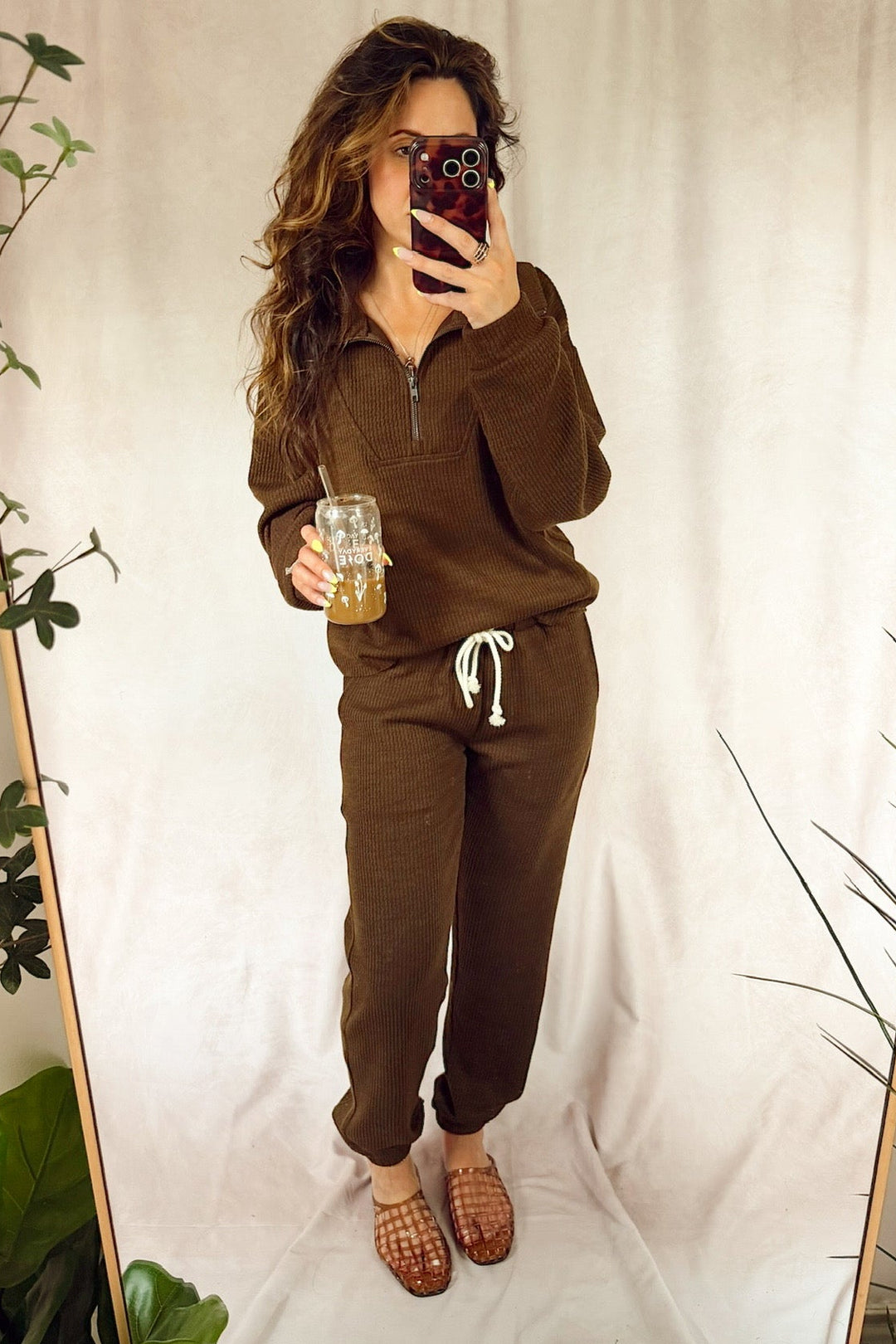 A woman takes a mirror selfie wearing a matching brown lounge set consisting of a half-zip pullover and jogger pants with a white drawstring. She is holding an iced coffee in front of a draped beige fabric backdrop with decorative plants on either side.