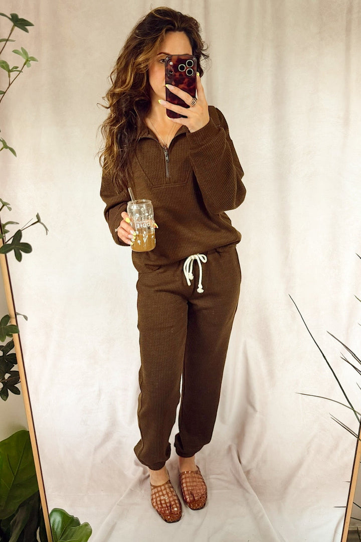 A woman takes a mirror selfie wearing a matching brown lounge set consisting of a half-zip pullover and jogger pants with a white drawstring. She is holding an iced coffee in front of a draped beige fabric backdrop with decorative plants on either side.